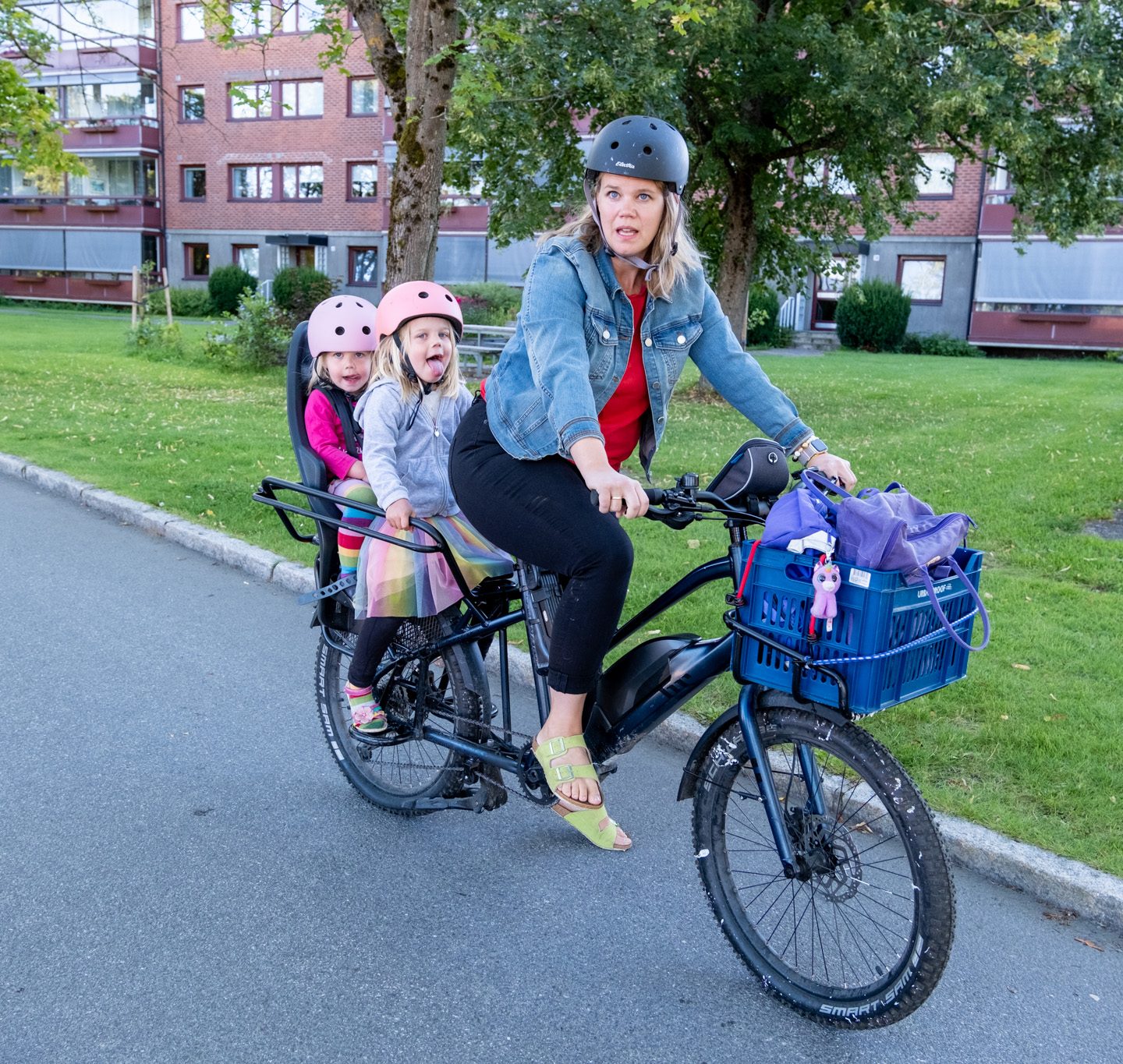 En kvinne kjører en lastesykkel med to barn bakerst, alle med hjelm. En kurv med poser er festet foran på sykkelen. Trær og bygninger er i bakgrunnen.