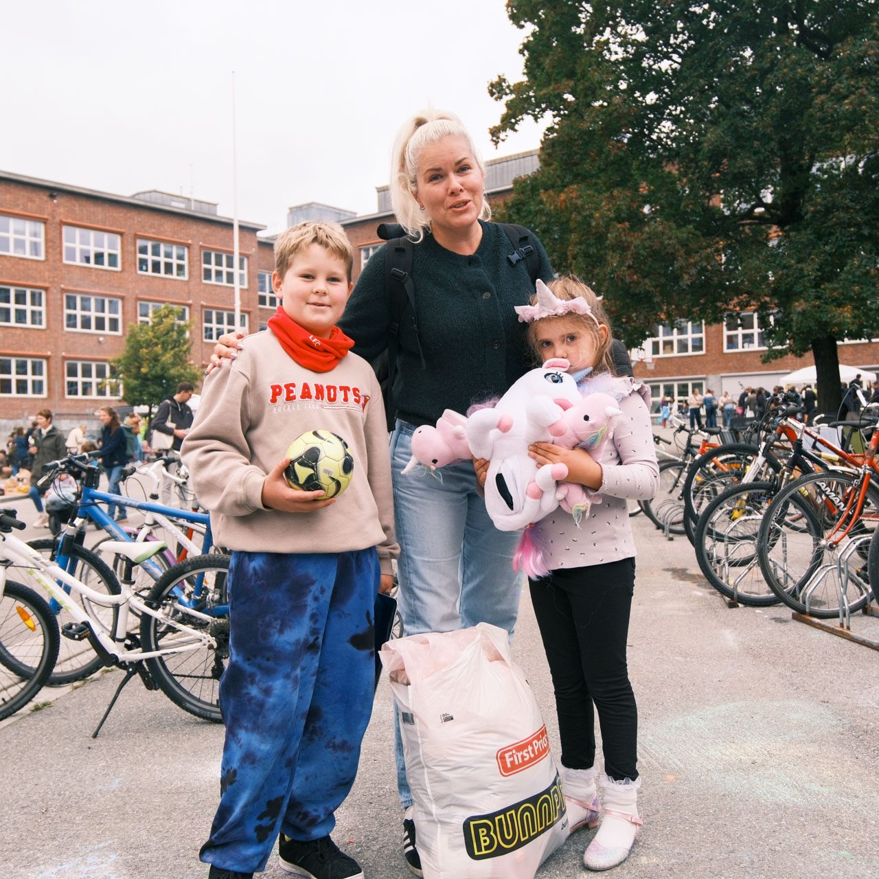 En kvinne og to barn står i nærheten av sykler foran en murbygning. Det ene barnet holder en fotball, og det andre er kledd i kostyme og holder en utstoppet leke.