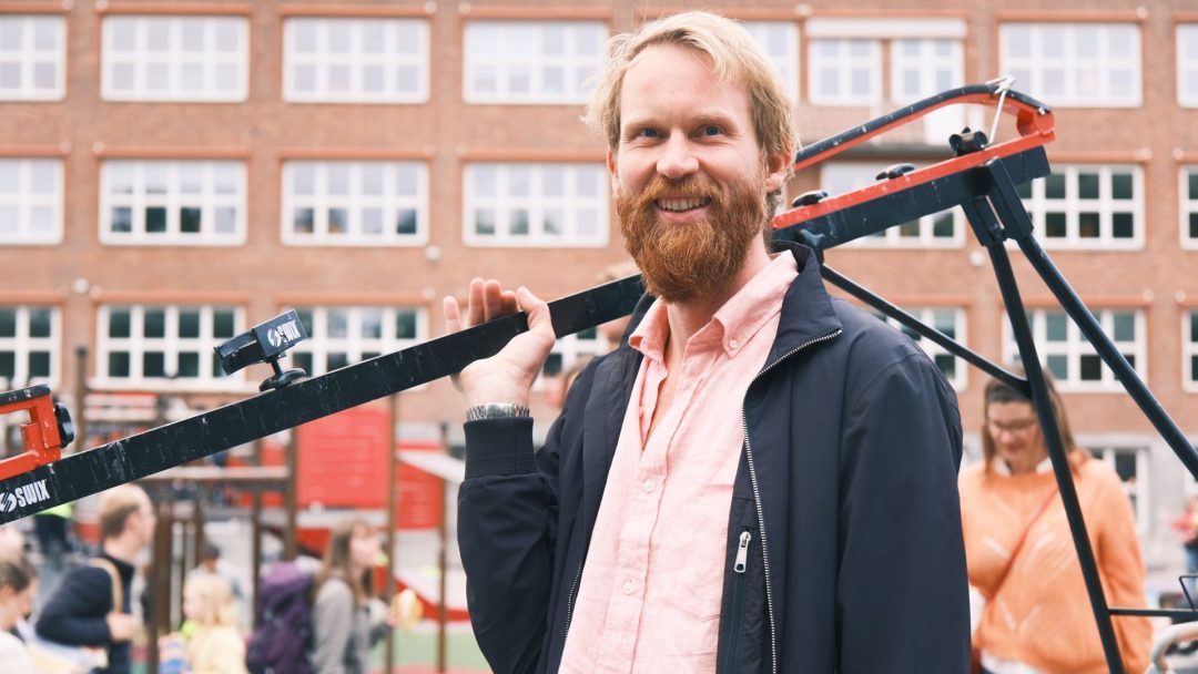 En skjeggete mann i rosa skjorte og jakke smiler mens han holder filmutstyr utendørs, med en murbygning og mennesker i bakgrunnen.