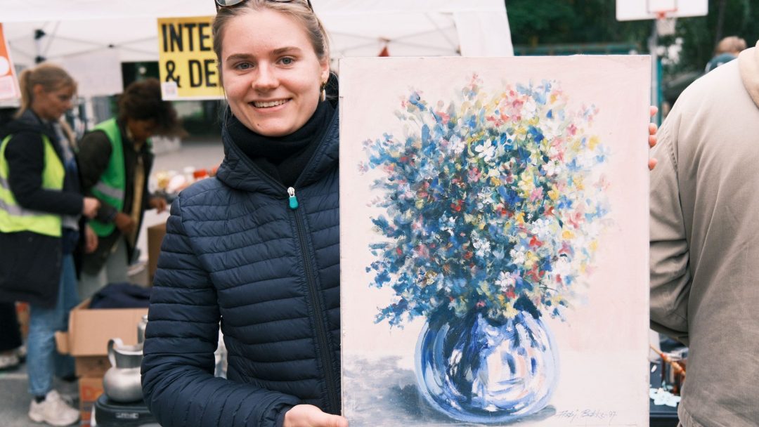 En person holder et fargerikt blomstermaleri i en vase på et utendørs arrangement. Folk og stander er synlige i bakgrunnen.