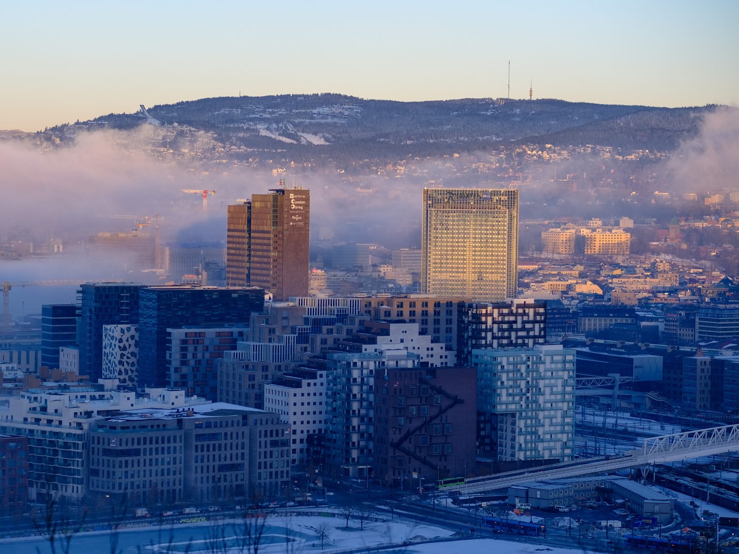 Luftfoto av et storbyer bybilde med moderne bygninger, en fjern tåkete horisont og et snødekt område i forgrunnen, som gjenspeiler gjennomtenkt planlegging innenfor klimabudsjettet.