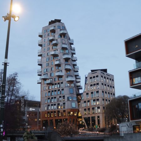 De moderne, unikt formede bygningene i Vertikal Nydalen med kantete design og mange vinduer står høyt mot en skumringshimmel. Gatelys lyser opp scenen, mens trær svaier forsiktig i forgrunnen.