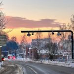 Snødekt byvei ved solnedgang med trafikkkameraer over hodet og bygninger i det fjerne. Elvarebiler glir stille langs gaten omkranset av nakne trær, og legger til et snev av modernitet til den rolige vinterscenen.