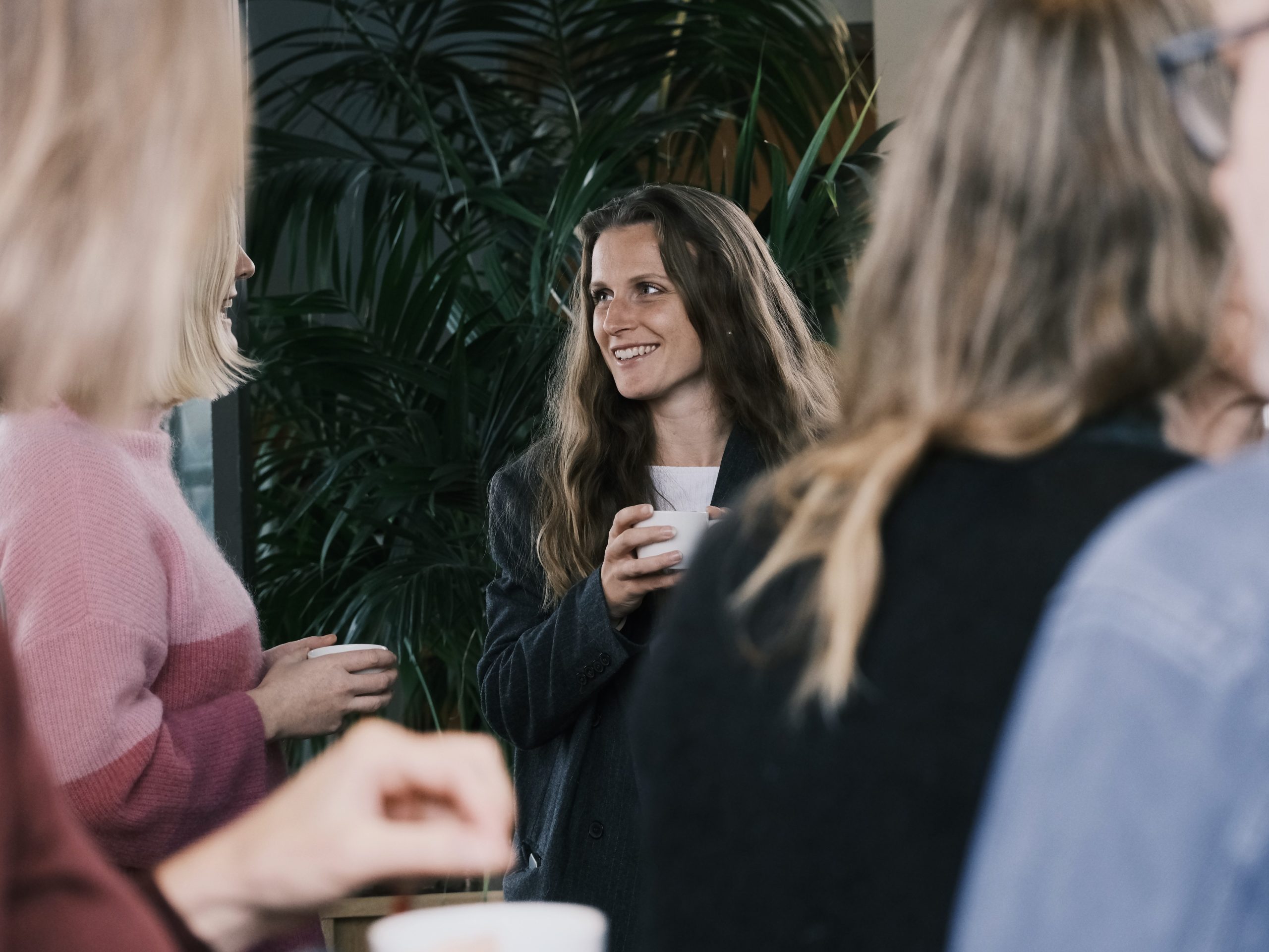 En gruppe mennesker som står innendørs, en kvinne i sentrum smiler og holder en kopp, med planter i bakgrunnen.