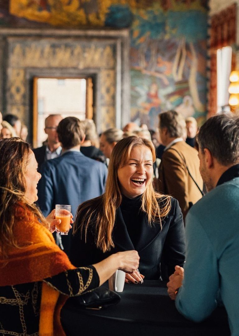 En gruppe mennesker sosialiserer seg på en sammenkomst, med en kvinne som ler og holder en drink. Fargerikt veggmaleri i bakgrunnen.