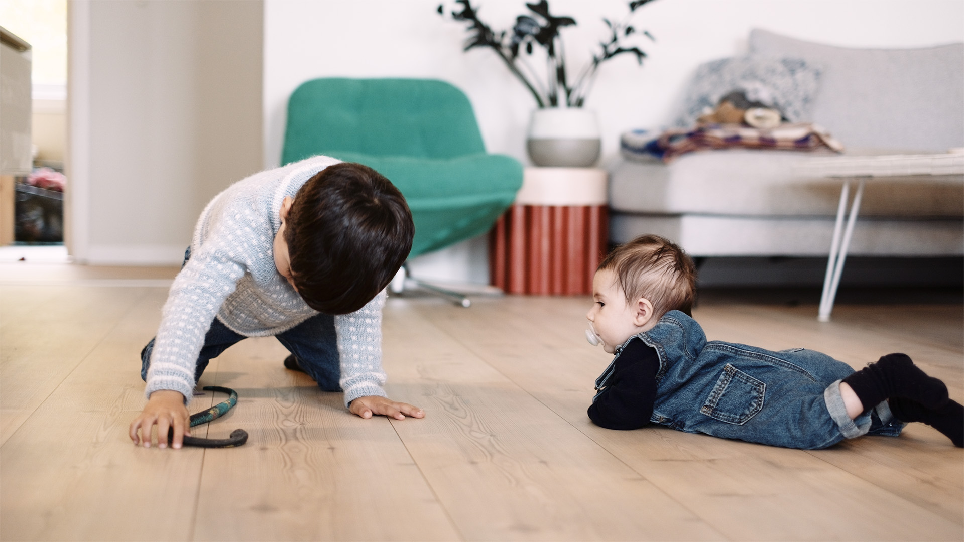 Et barn kryper på gulvet, fascinert av et stetoskop, mens en baby hviler på magen i nærheten. Stuen, med en grønn stol og sofa, lyses opp av energieffektive vinduer og ytterdører.