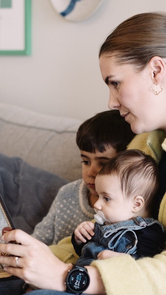 En kvinne som sitter på en sofa holder en smarttelefon, med to barn, ett eldre og en baby, plassert tett ved siden av henne. Mens hun utforsker nye muligheter på enheten sin, følger deres nysgjerrige øyne med, betatt av de uendelige mulighetene.