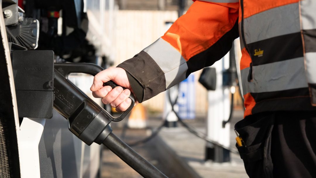 Person i en oransje sikkerhetsjakke som holder en ladeplugg for elektrisk kjøretøy, forbereder seg på å lade et kjøretøy.