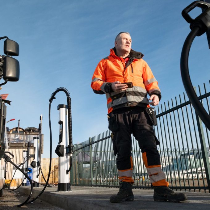 Person i en oransje sikkerhetsjakke står nær elektriske ladestasjoner og lastebiler, holder en enhet, med klar himmel og inngjerdet område i bakgrunnen.