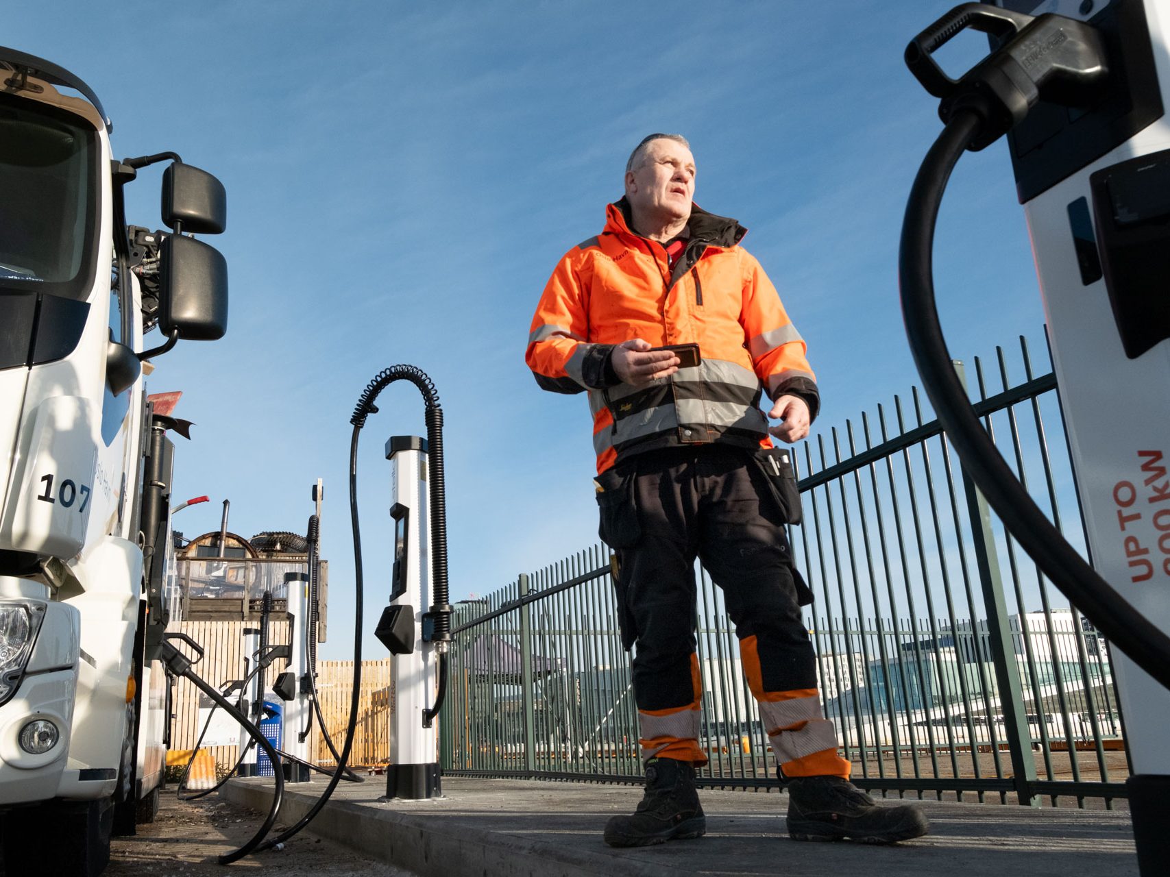 Person i en oransje sikkerhetsjakke står nær elektriske ladestasjoner og lastebiler, holder en enhet, med klar himmel og inngjerdet område i bakgrunnen.