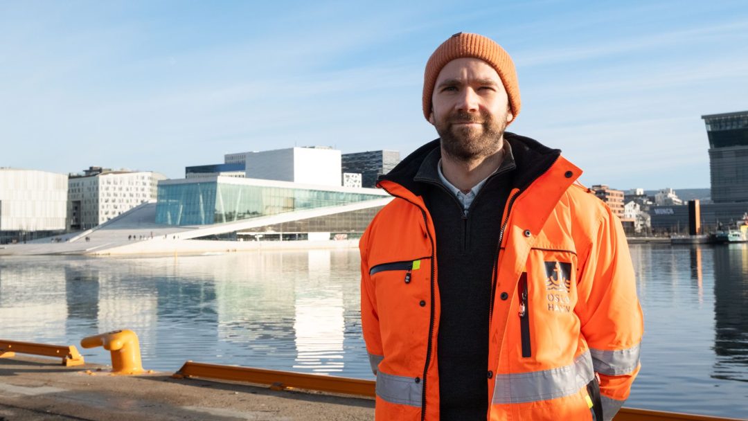 En person i oransje jakke og lue står ved vannkanten med moderne bygninger og klar himmel i bakgrunnen.