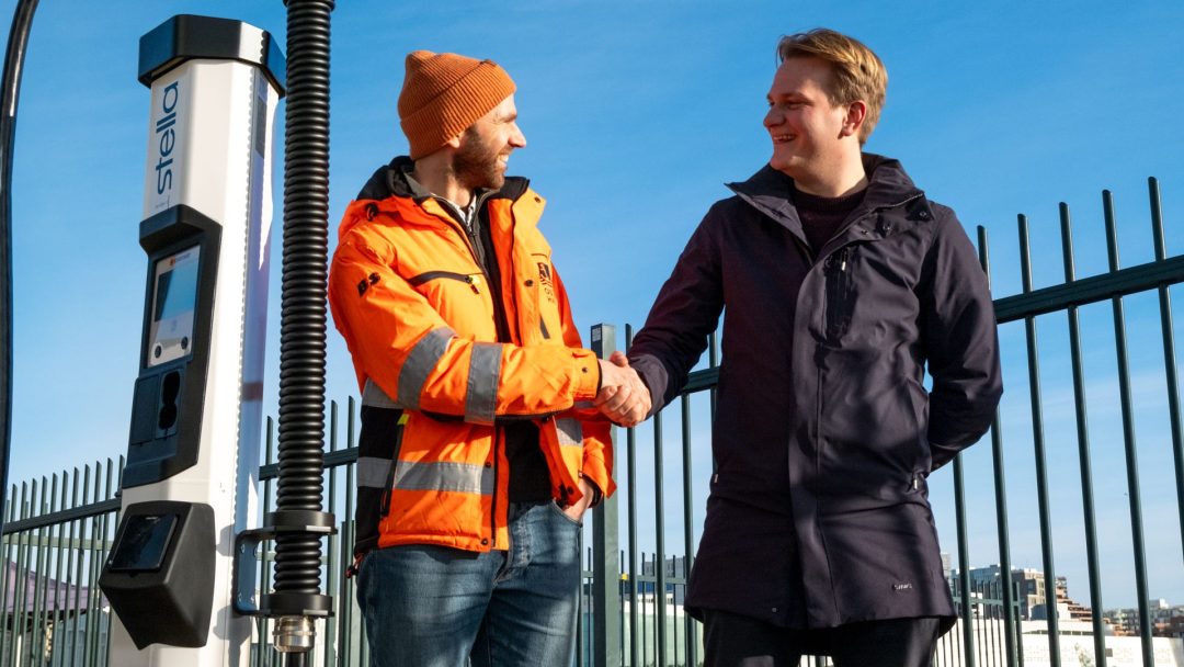 To menn håndhilser nær en ladestasjon for elektriske kjøretøy på en solrik dag. Den ene har på seg en oransje jakke og lue, den andre en mørk frakk. Et metallgjerde og bybilde er i bakgrunnen.