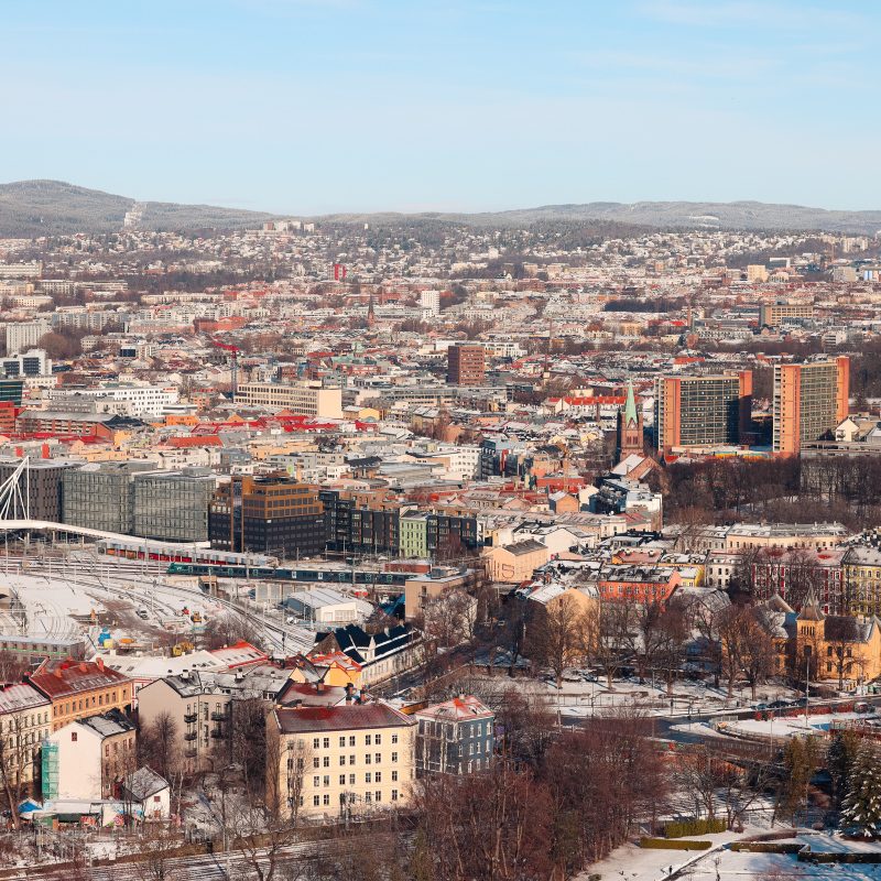 En luftfoto av et bybilde avslører mange bygninger, veier og bladløse trær under en klar himmel. Høye strukturer står langs fjerne åser mens snøflekker sprer landskapet, og minner innbyggerne om å sparre strøm under den raske vinterkulden.