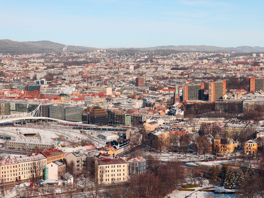 En energikart fanger et luftbilde av et bybilde med tette bygninger, en bro og omkringliggende åser. Snø tepper deler av bakken og hustak under en klar blå himmel.