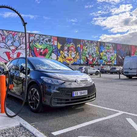 Svart Volkswagen ID.3 lader på en stasjon på en parkeringsplass. Et fargerikt veggmaleri med tegneseriefigurer er synlig i bakgrunnen under en delvis overskyet himmel.