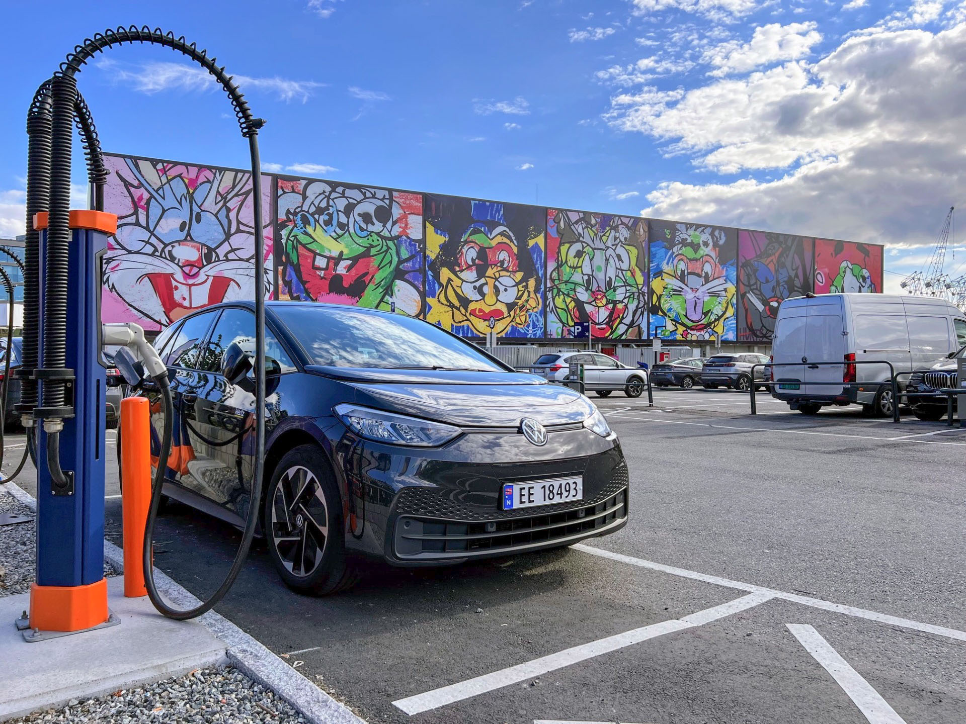 Svart Volkswagen ID.3 lader på en stasjon på en parkeringsplass. Et fargerikt veggmaleri med tegneseriefigurer er synlig i bakgrunnen under en delvis overskyet himmel.