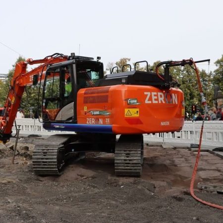 En oransje gravemaskin merket ZERON takler byggeplassen, omgitt av steinsprut og maskiner, som minner om nyvinninger diskutert på energikonferanser. Trær og bygninger står vakt i bakgrunnen.