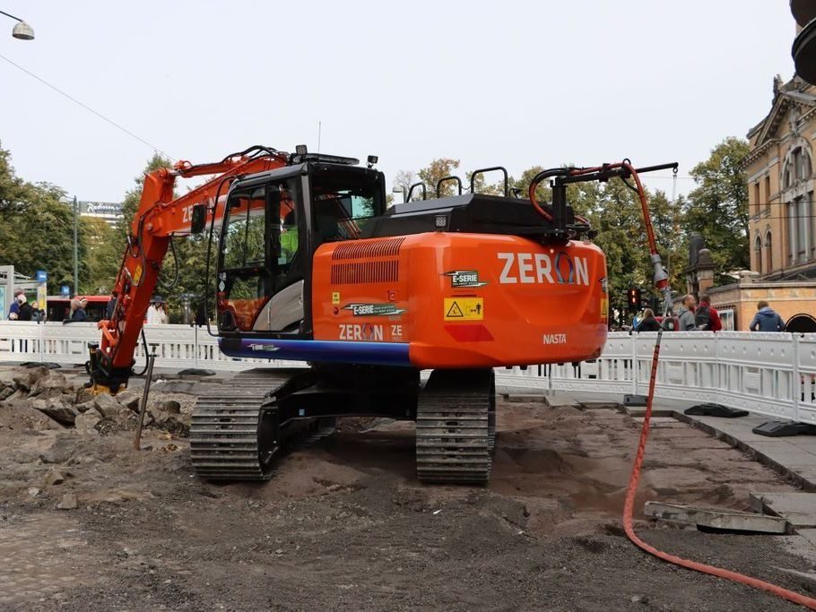 En oransje gravemaskin merket ZERON takler byggeplassen, omgitt av steinsprut og maskiner, som minner om nyvinninger diskutert på energikonferanser. Trær og bygninger står vakt i bakgrunnen.