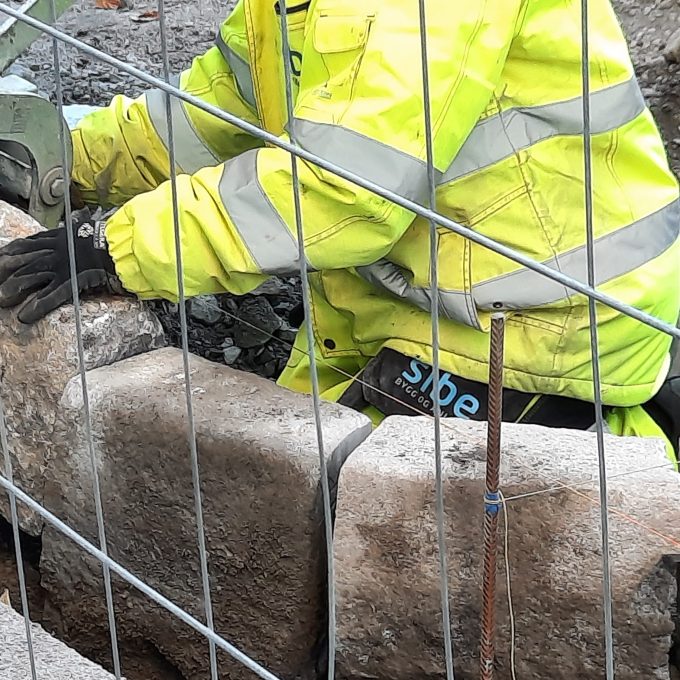 En arbeider i en synlig jakke manøvrerer omhyggelig store Stein-steinblokker på en byggeplass, alt trygt observert bak et solid metallgjerde.